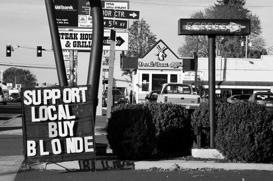 "Support Local" reads a sign in front of Blondie's Coffee stand. Blondie's is a locally owned, one of a kind espresso shop. In the background you can see the presence of the corporate monopoly called Dutch Bros. To date, there are 137 Dutch Bros. franchises.
